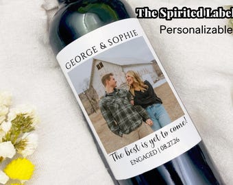 May include: A white wine bottle label featuring a photograph of a couple in front of a barn. The label text reads "George & Sophie" and "The best is yet to come! Engaged | 08.27.26".