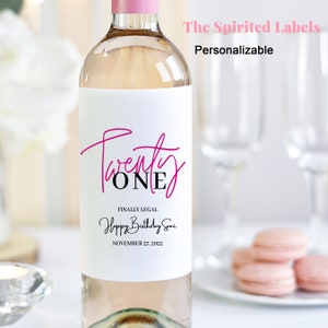 May include: A white wine bottle with a personalised label that says "Twenty One" in pink and black lettering. The label also says "Finally Legal Happy Birthday Somi November 27, 2022".
