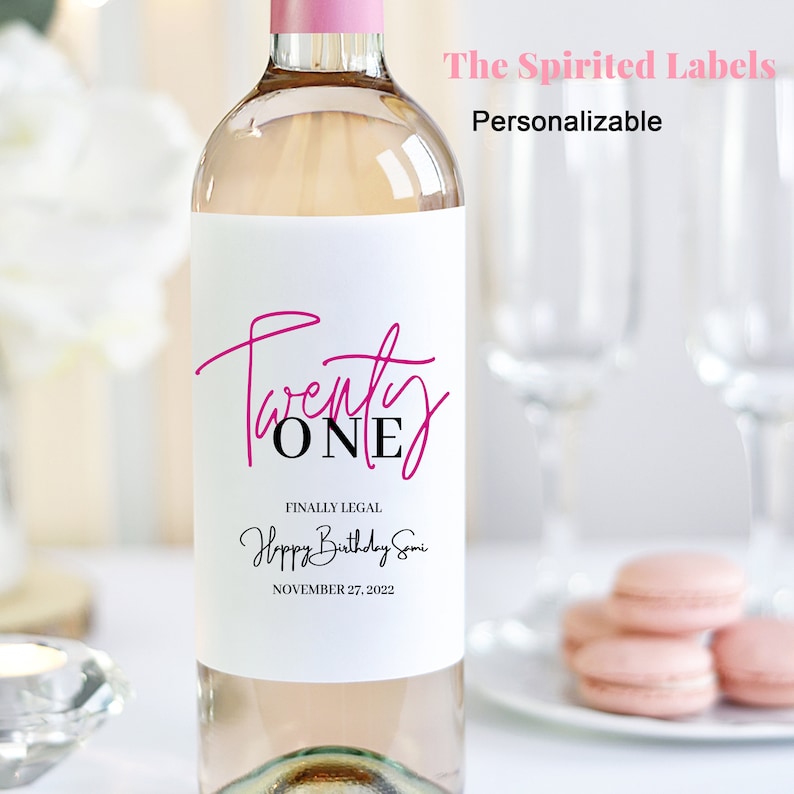 May include: A white wine bottle with a personalised label that says "Twenty One" in pink and black lettering. The label also says "Finally Legal Happy Birthday Somi November 27, 2022".