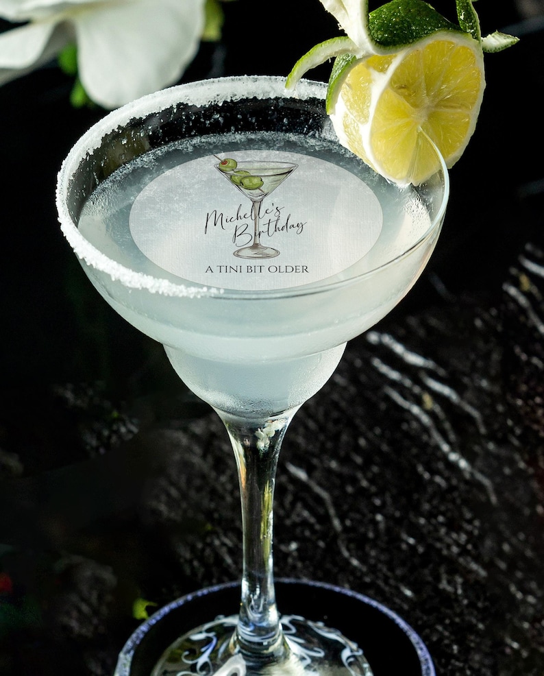 May include: A white margarita glass with a salted rim, filled with a clear liquid and a green olive garnish. A round sticker on the glass reads "Michelle's Birthday A TINI BIT OLDER".