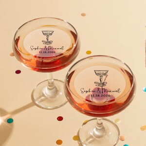 May include: Two champagne flutes with personalized stickers. The stickers are white with black text and a drawing of a champagne glass. The text on the stickers reads "Sophia & Manuel 11.18.2026".