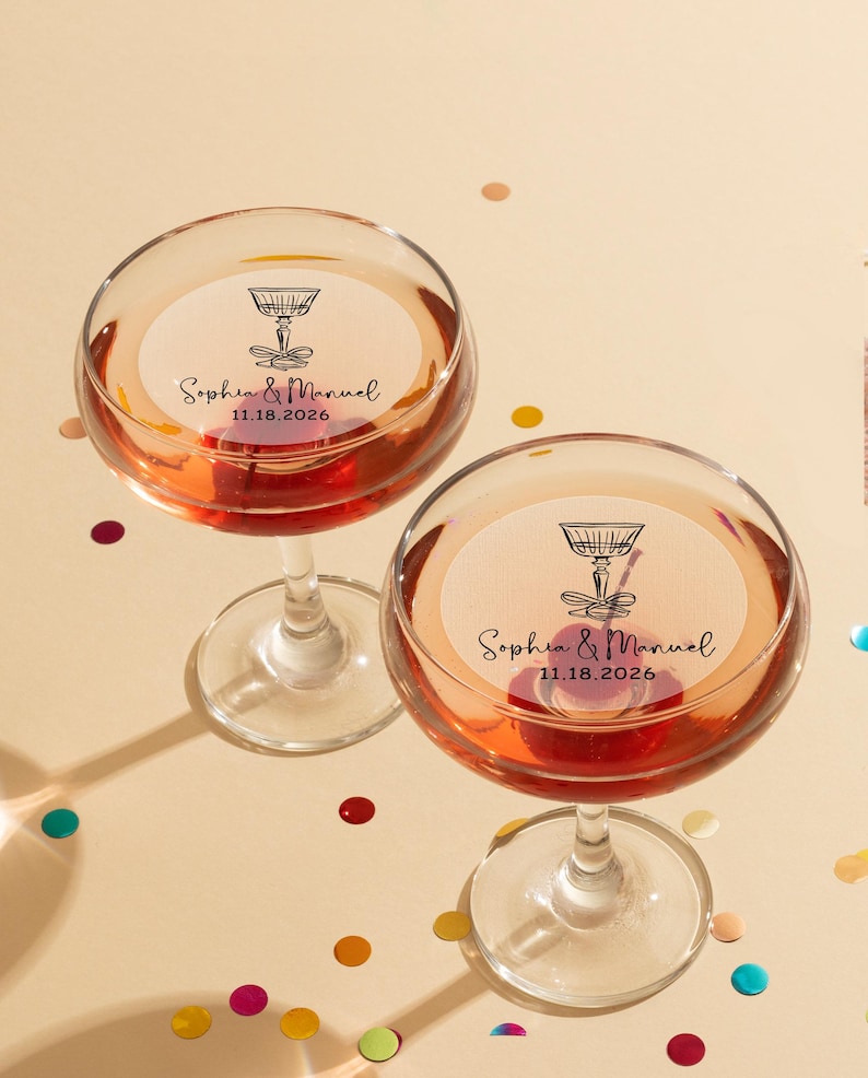 May include: Two champagne flutes with personalized stickers. The stickers are white with black text and a drawing of a champagne glass. The text on the stickers reads "Sophia & Manuel 11.18.2026".