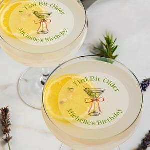 May include: Two martini glasses with a lemon slice and a sticker that says "A Tini Bit Older Michelle's Birthday" with a drawing of a martini glass with olives.