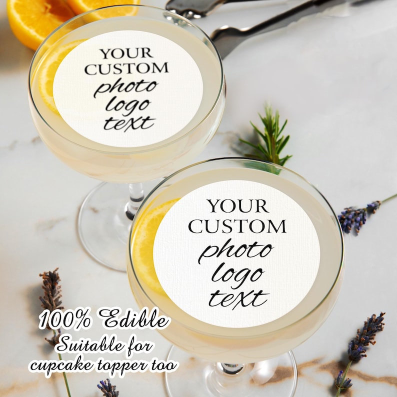 May include: Two clear glass cocktail glasses with a white round edible sticker on each. The stickers have black text that says "YOUR CUSTOM photo logo text". The text is written in a cursive font. The glasses are filled with a light yellow liquid. The text "100% Edible Suitable for cupcake topper too" is written below the glasses.