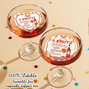 May include: Two stemmed glasses filled with a reddish-orange liquid, each topped with a heart-shaped edible topper. The toppers read "A Spritz, Older, Happy Birthday Olivia" with a graphic of a cocktail and a bottle. The text "100% Edible, Suitable for cupcake topper too" is at the bottom.