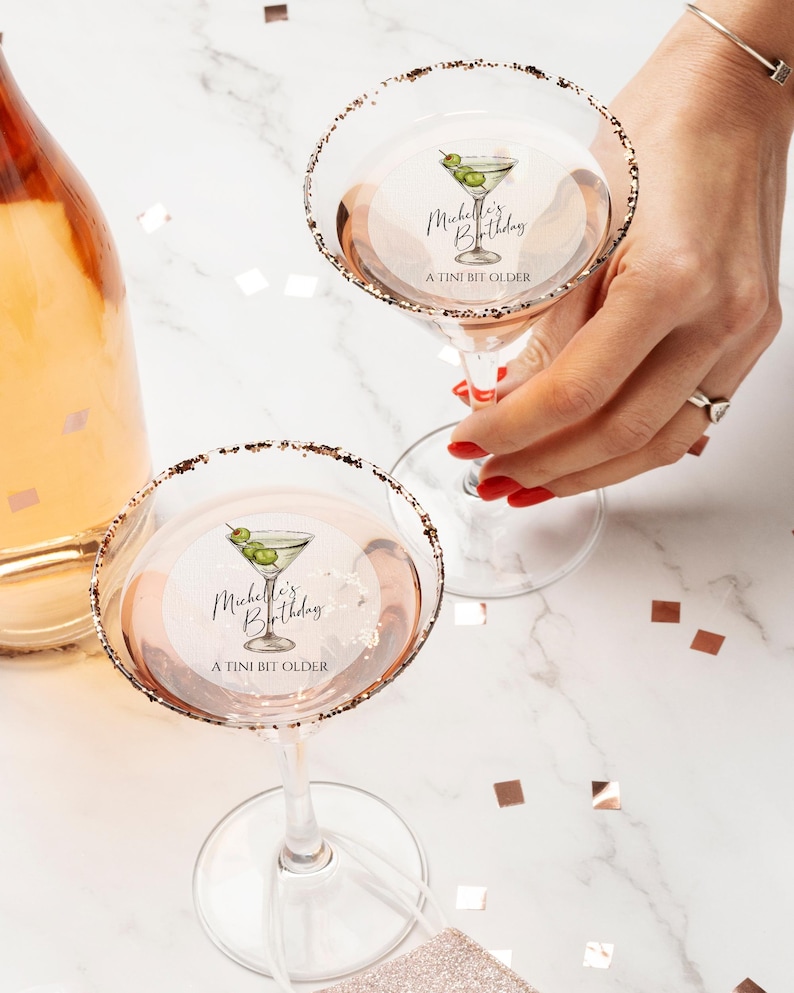 May include: Two martini glasses with gold rims and a personalized sticker that says "Michelle's Birthday A TINI BIT OLDER" with a drawing of a martini glass with olives.