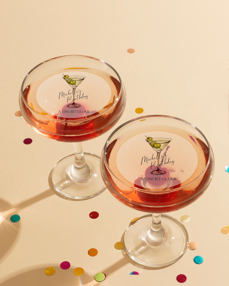 May include: Two martini glasses filled with a pink beverage. Each glass has a round sticker with a martini glass illustration and the text "Michelle's Birthday A TINI BIT OLDER".