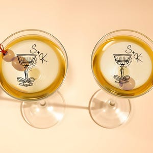 May include: Two clear glass martini glasses with gold rims. Each glass has a black and white sticker with a stylized illustration of a martini glass and the text "S+K 11.20.2026".