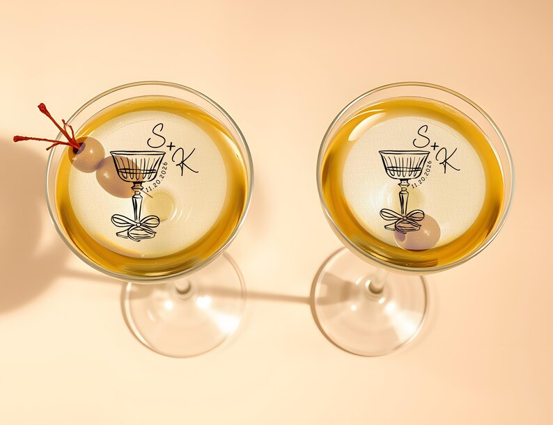 May include: Two clear glass martini glasses with gold rims. Each glass has a black and white sticker with a stylized illustration of a martini glass and the text "S+K 11.20.2026".