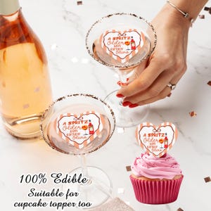 May include: A bottle of sparkling wine and two martini glasses with sugared rims. Each glass has a heart-shaped topper that reads "A Spritz! Older Happy Birthday Olivia." A cupcake with a matching topper is also present. The text "100% Edible Suitable for cupcake topper too" is at the bottom.