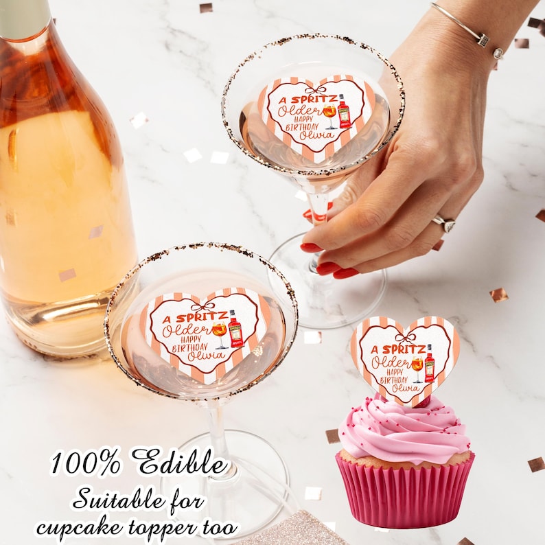May include: A bottle of sparkling wine and two martini glasses with sugared rims. Each glass has a heart-shaped topper that reads "A Spritz! Older Happy Birthday Olivia." A cupcake with a matching topper is also present. The text "100% Edible Suitable for cupcake topper too" is at the bottom.