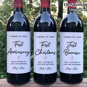 May include: Three wine bottles with personalized labels. The labels are white with black text and a pink heart. The text on the labels reads "Cheers to your First Anniversary", "Cheers to your First Christmas", and "Cheers to your Just Because". Each label also includes the text "And a lifetime of love and happiness" followed by the names "Jenny & Chris".