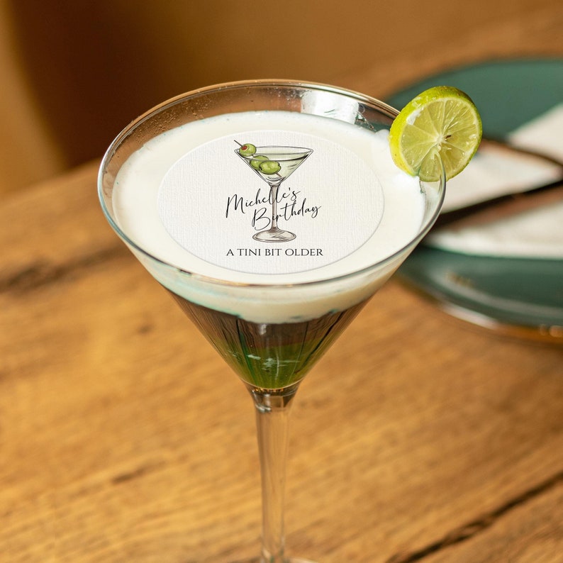 May include: A white round paper coaster with a martini glass illustration and the text "Michelle's Birthday A TINI BIT OLDER". The coaster is on top of a martini glass filled with a layered green, white, and brown drink. A lime wedge is on the rim of the glass.