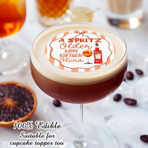 May include: A cocktail in a stemmed glass with a white foam top, featuring an edible topper. The topper has the text "A Spritz Older Happy Birthday Olivia" with illustrations of a cocktail and an Aperol bottle. The image also includes coffee beans and a slice of orange.