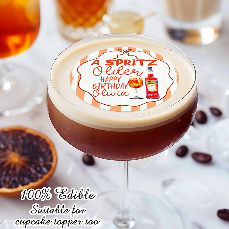 May include: A cocktail in a stemmed glass with a white foam top, featuring an edible topper. The topper has the text "A Spritz Older Happy Birthday Olivia" with illustrations of a cocktail and an Aperol bottle. The image also includes coffee beans and a slice of orange.