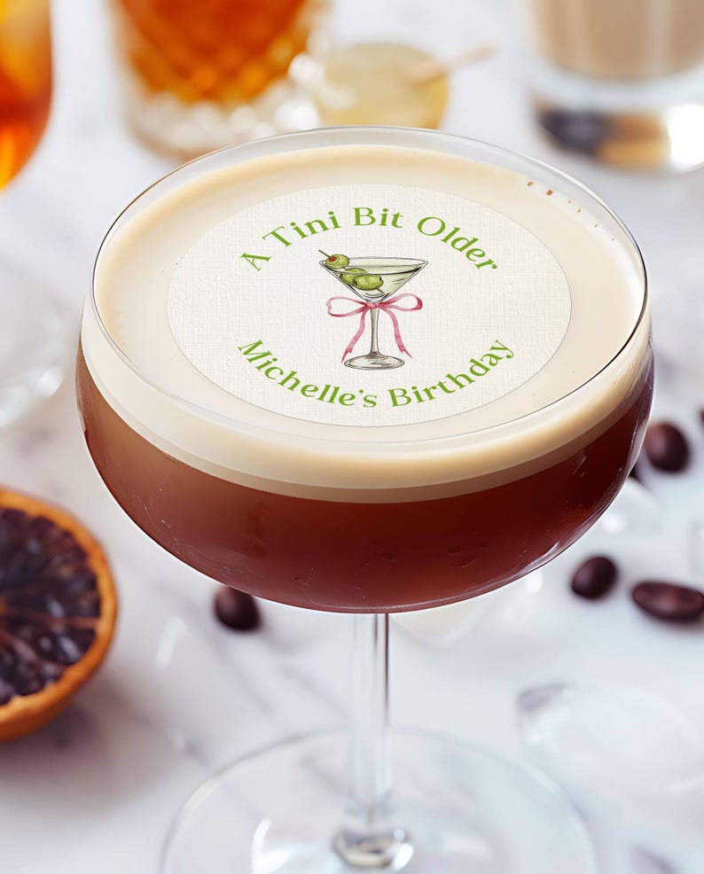 May include: A white round sticker with green text that reads "A Tini Bit Older Michelle's Birthday". The sticker has a green illustration of a martini glass with olives and a pink ribbon.