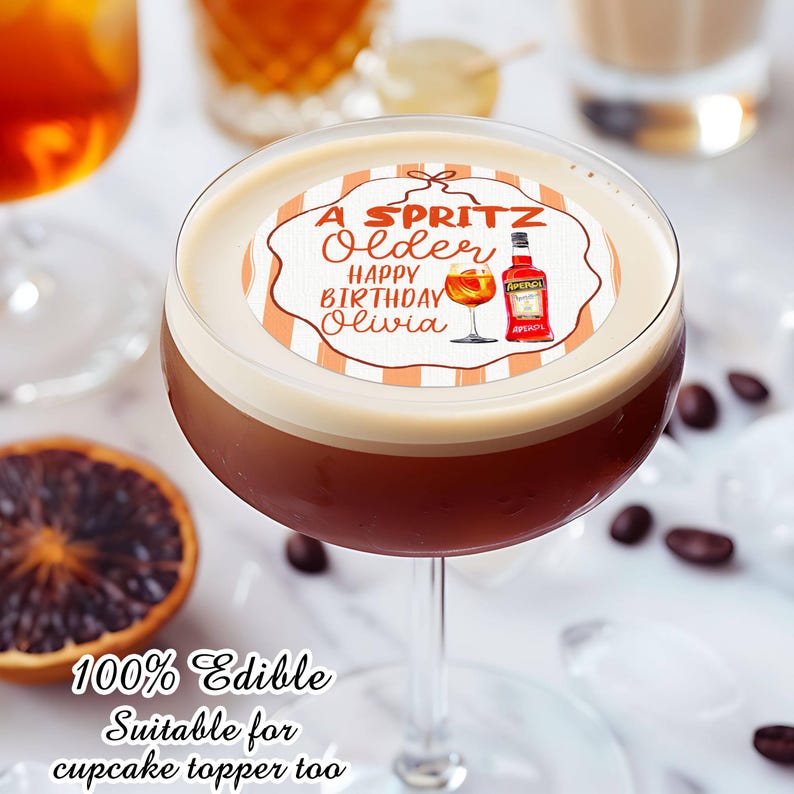 May include: A cocktail in a stemmed glass with a white foam top, featuring an edible topper. The topper has the text "A Spritz Older Happy Birthday Olivia" with illustrations of a cocktail and an Aperol bottle. The image also includes coffee beans and a slice of orange.