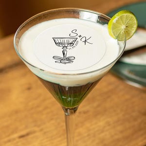 May include: A green and white cocktail in a martini glass with a white round paper coaster on top. The coaster has a black illustration of a champagne glass with a bow and the text "S+K 11.20.2026".