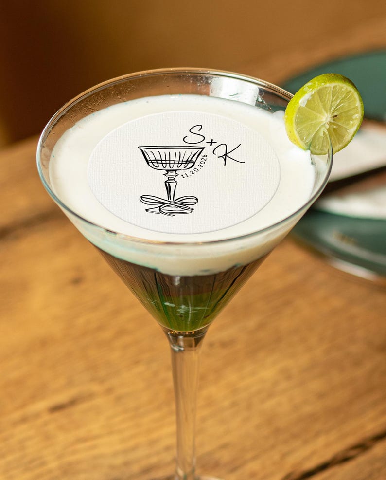 May include: A green and white cocktail in a martini glass with a white round paper coaster on top. The coaster has a black illustration of a champagne glass with a bow and the text "S+K 11.20.2026".