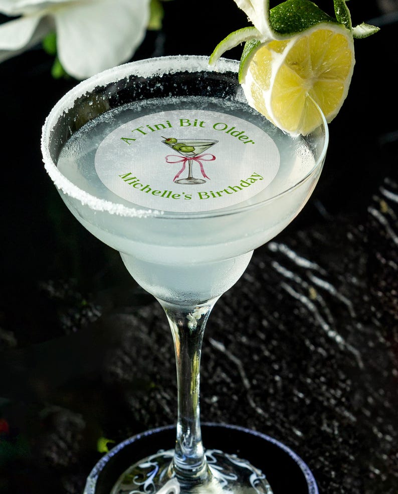 May include: A round, white, paper cocktail napkin with green text that reads "A Tini Bit Older Michelle's Birthday" and a green illustration of a martini glass with olives. The napkin is placed on top of a margarita glass filled with a clear liquid and a lime wedge.