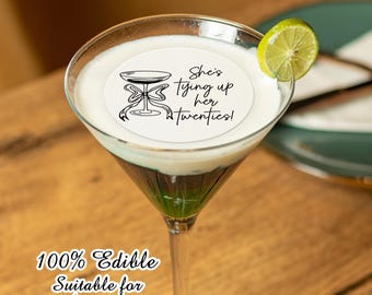 May include: A round edible drink topper featuring a black line drawing of a martini glass with a bow and the text She's tying up her twenties. The white topper floats on a creamy cocktail garnished with a fresh green lime slice.