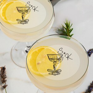 May include: Two clear glass cocktail glasses with a lemon slice garnish. Each glass has a white round sticker with a black illustration of a cocktail glass and the text "S+K 11.20.2026".