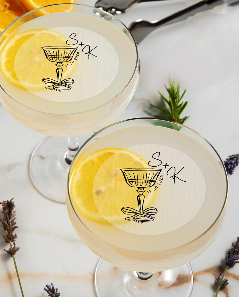 May include: Two clear glass cocktail glasses with a lemon slice garnish. Each glass has a white round sticker with a black illustration of a cocktail glass and the text "S+K 11.20.2026".