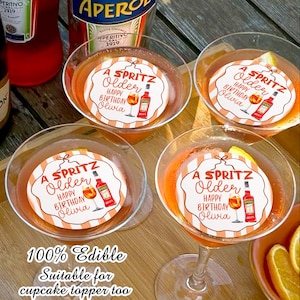 May include: Four martini glasses filled with an orange cocktail, each topped with a circular edible image. The images read "A Spritz Older Happy Birthday Olivia" with a bottle and glass illustration. The text "100% Edible Suitable for cupcake topper too" is at the bottom.