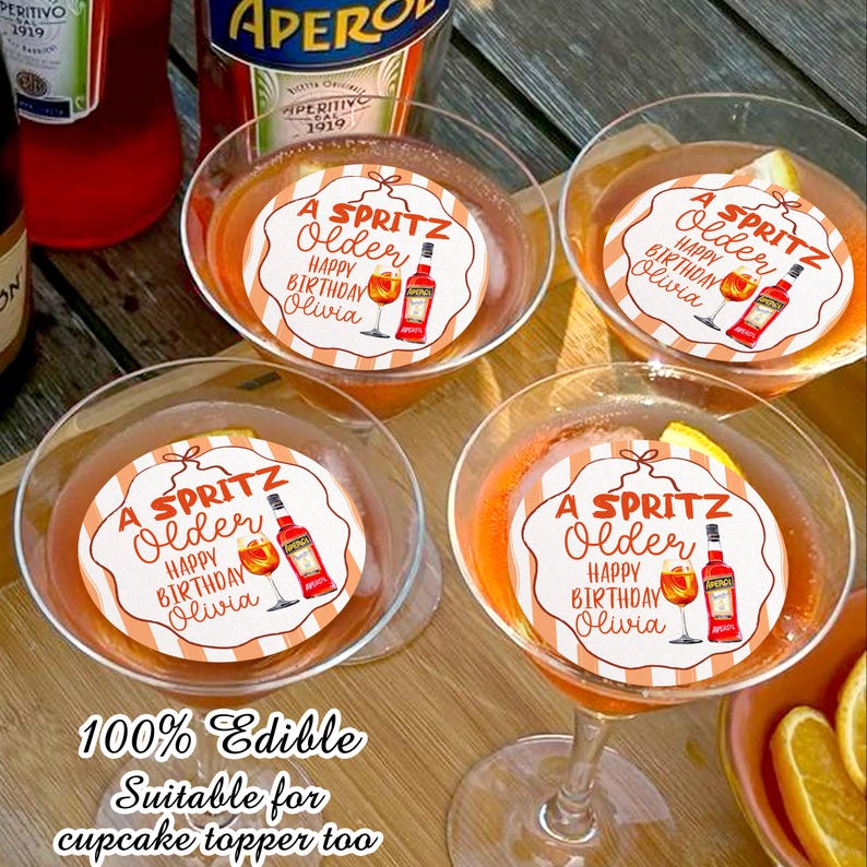 May include: Four martini glasses filled with an orange cocktail, each topped with a circular edible image. The images read "A Spritz Older Happy Birthday Olivia" with a bottle and glass illustration. The text "100% Edible Suitable for cupcake topper too" is at the bottom.