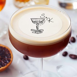 May include: A close-up of a cocktail with a white foam top and a round, white paper coaster with black ink. The coaster has a drawing of a stemmed glass with a bow and the text "S+K 11.20.2020".