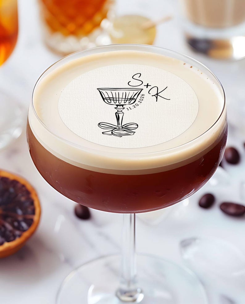 May include: A close-up of a cocktail with a white foam top and a round, white paper coaster with black ink. The coaster has a drawing of a stemmed glass with a bow and the text "S+K 11.20.2020".