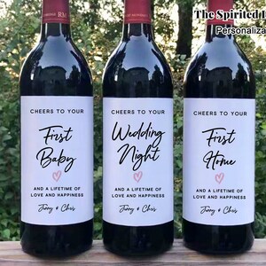 May include: Three wine bottles with personalized labels. The labels are white with black text and a pink heart. The text on the labels reads "Cheers to your First Baby", "Cheers to your Wedding Night", and "Cheers to your First Home".