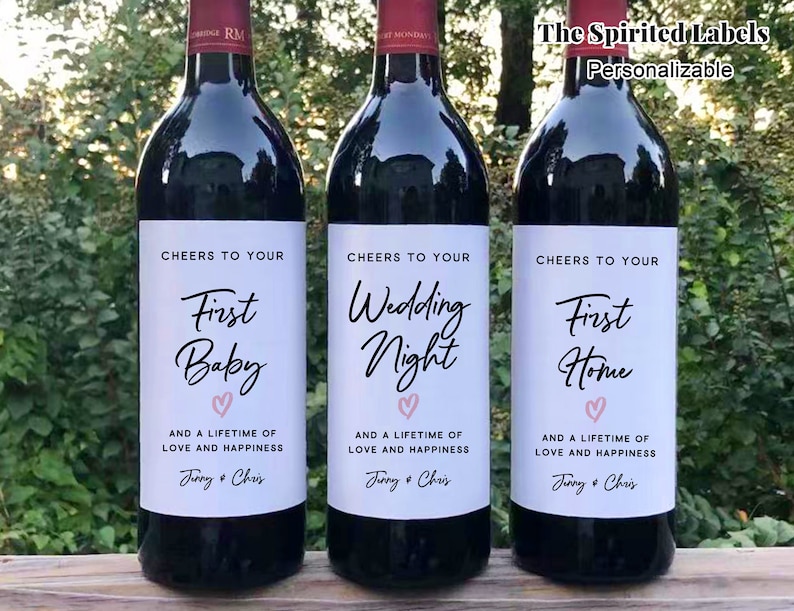 May include: Three wine bottles with personalized labels. The labels are white with black text and a pink heart. The text on the labels reads "Cheers to your First Baby", "Cheers to your Wedding Night", and "Cheers to your First Home".