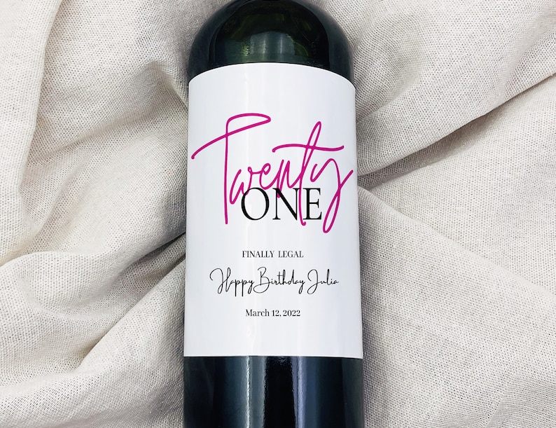 May include: A wine bottle with a white label that says "Twenty ONE" in pink script and "FINALLY LEGAL" in black text. Below that, it says "Happy Birthday Julie" in black script and "March 12, 2022" in black text.