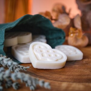 Può includere: Una borsa di stoffa verde piena di saponi bianchi a forma di cuore con un design a nodo celtico. I saponi sono su una superficie di legno con lavanda secca e bacche rosse.
