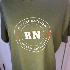 May include: A green t-shirt with a white circle graphic that says "A Little Ratched A Little Nightingale" with a red heartbeat and the letters "RN" in the center.