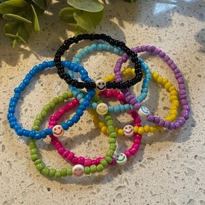 May include: A set of colorful beaded bracelets with smiley face charms. The bracelets are made with small, round beads in various colors, including black, blue, green, yellow, and purple. Each bracelet has a single white smiley face charm.