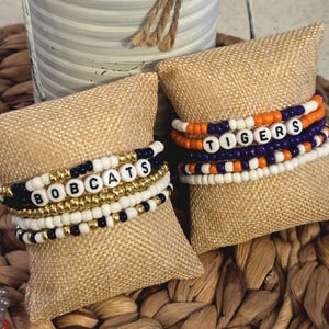 May include: Stacked beaded bracelets with the words "BOBCATS" and "TIGERS" spelled out in white letter beads. The bracelets feature gold, white, navy blue, and orange beads. They are displayed on burlap-covered pillows.