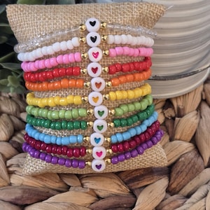 May include: A stack of colorful beaded bracelets with heart-shaped charms. Each bracelet is a different color, including white, pink, orange, yellow, green, blue, red, and purple. The charms are white with a colored heart in the center.