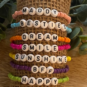 May include: A stack of colorful beaded bracelets with different words spelled out on them, including "Maddy", "Besties", "Dream", "Smile", "Sunshine", "BFF", and "Happy".