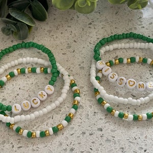 May include: Set of four beaded bracelets with green and white beads. One bracelet has the word "LUCKY" spelled out in white beads with gold lettering. The other bracelet has the word "KISS" spelled out in white beads with gold lettering, followed by a heart bead and the word "ME" spelled out in white beads with gold lettering.