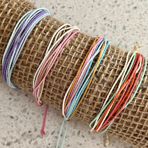 May include: A set of five colorful friendship bracelets made with thin, braided cords. The bracelets are in shades of pink, blue, orange, and purple. The bracelets are wrapped around a piece of burlap fabric.