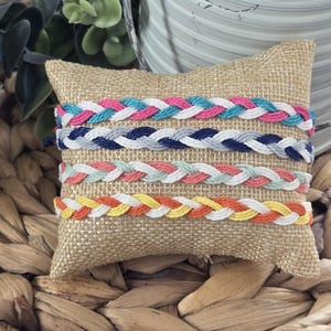 May include: Braided friendship bracelets in various colors, including pink, blue, gray, and yellow, are displayed on a burlap pillow. The bracelets are made of woven threads and are presented on a woven wicker surface.