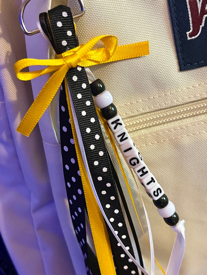 K&ouml;nnte beinhalten: Ein Schl&uuml;sselband aus schwarz-wei&szlig;em Polka-Dot-Band mit einer gelben Schleife und wei&szlig;en Perlen, auf denen das Wort "KNIGHTS" in schwarzen Buchstaben steht.