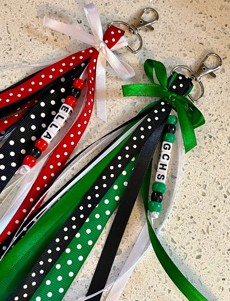K&ouml;nnte beinhalten: Zwei Schl&uuml;sselanh&auml;nger mit schwarz-wei&szlig;em Polka-Dot-Band, wei&szlig;em Band und gr&uuml;nem Band. Ein Schl&uuml;sselanh&auml;nger hat den Namen "Ella" in wei&szlig;en Perlen ausgeschrieben und der andere die Buchstaben "GCHS" in wei&szlig;en Perlen.