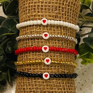 May include: Five bracelets with red heart charms on a burlap background. The bracelets are white, silver, red, gold, and black.