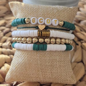May include: Stack of four bracelets with green, white, and gold beads. One bracelet spells out the letters "G C D T" with gold letter beads. A small gold heart charm is also included.