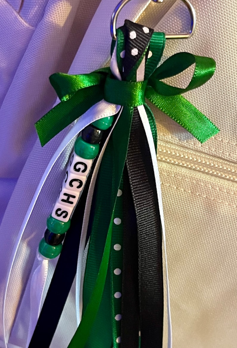 K&ouml;nnte beinhalten: Ein gr&uuml;ner, schwarzer und wei&szlig;er Schl&uuml;sselband mit einem Perlenanh&auml;nger, der "GCHS" buchstabiert.