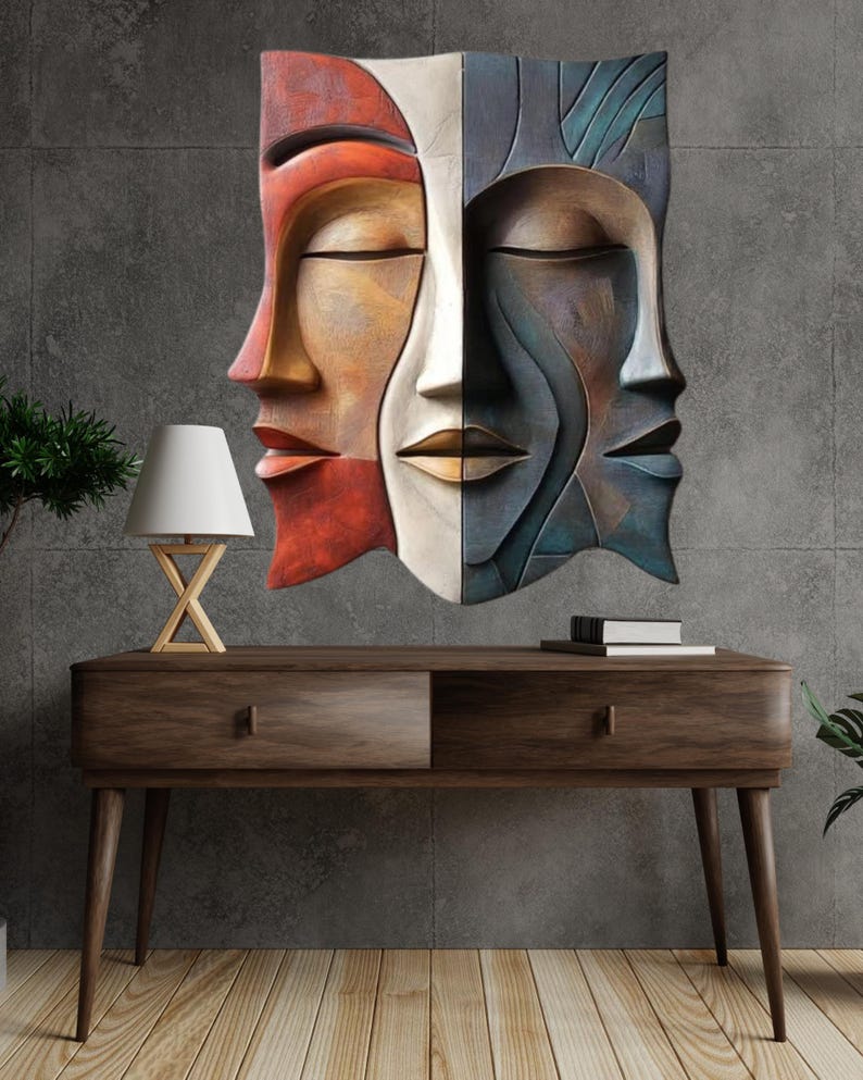May include: A multi-colored wall sculpture depicting three stylized faces with closed eyes. The sculpture is mounted on a gray wall above a wooden console table. The faces are in shades of red, orange, white, and blue.