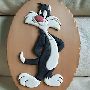 Puede incluir: Un adorno de pared de madera con Silvestre el Gato de Looney Tunes. El gato de dibujos animados está representado en negro, blanco y rojo, de pie sobre una placa ovalada marrón. La placa tiene un borde decorativo y un agujero para colgar.